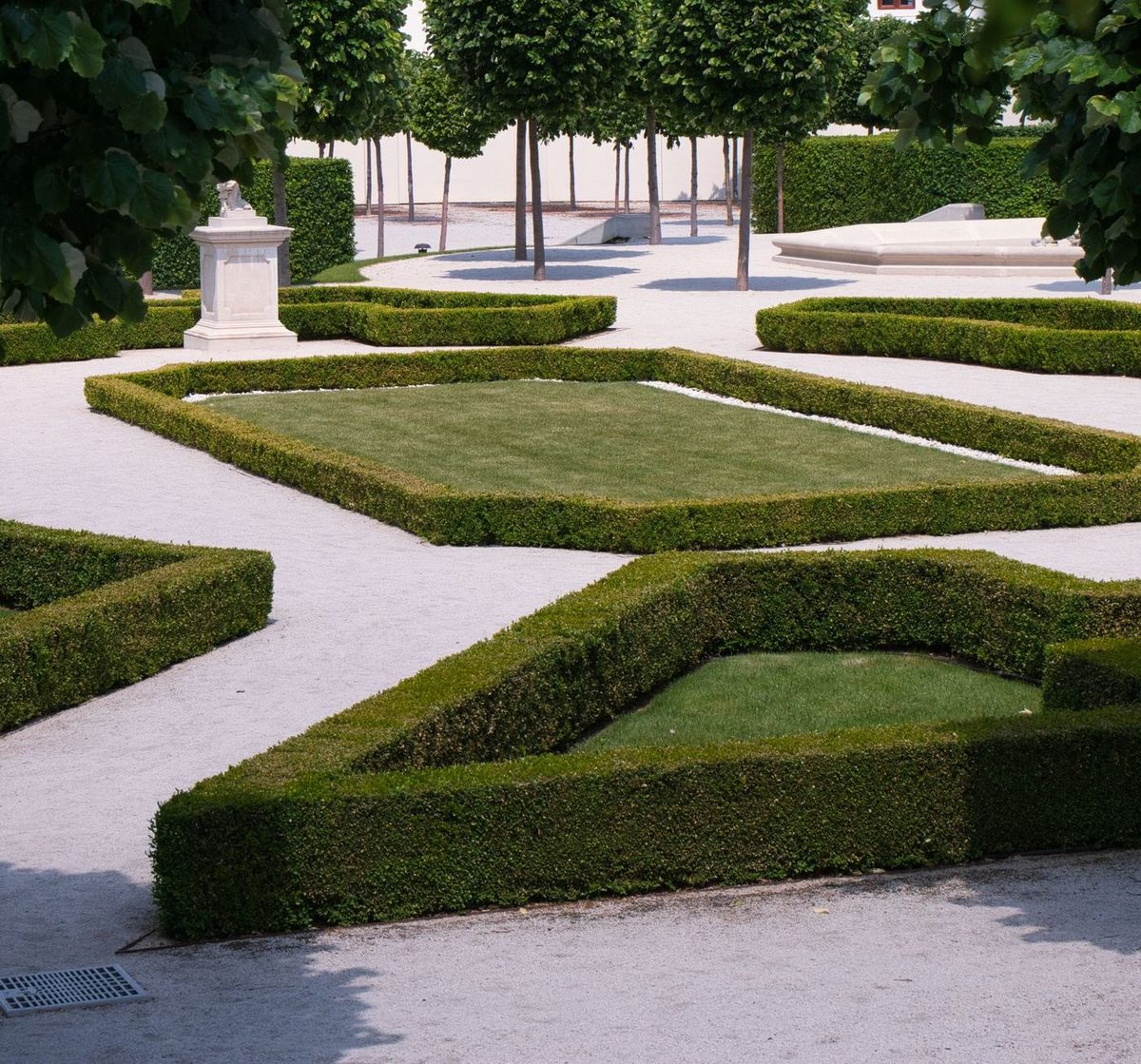 A formal parterre of low clipped box hedges enclosing lawn panels and white gravel, with a stone pedestal and a row of clipped lime trees